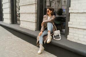 Outdoor autumn photo of young elegant fashionable woman wearing trendy sunglasses,  beige color coat, white textured leather shoulder bag, walking in street of European city.