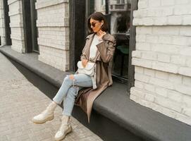 Elegant fashionable woman with short bruneete hairs  wearing trendy sunglasses,  beige color coat, white textured leather shoulder bag, walking in street of European city. photo