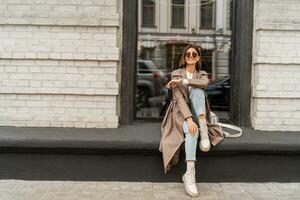 Outdoor autumn photo of young elegant fashionable woman wearing trendy sunglasses,  beige color coat, white textured leather shoulder bag, walking in street of European city.