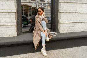 Outdoor autumn photo of young elegant fashionable woman wearing trendy sunglasses,  beige color coat, white textured leather shoulder bag, walking in street of European city.