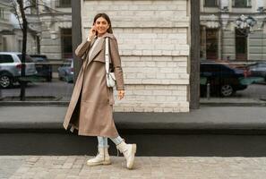 Confident short haired woman wearing  casual   beige color coat, white textured leather shoulder bag, walking in street of European city.  Autumn fashion trends.  Full lenght. photo