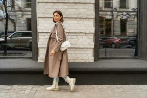 Confident short haired woman wearing  casual   beige color coat, white textured leather shoulder bag, walking in street of European city.  Autumn fashion trends.  Full lenght. photo