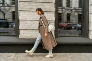 Confident short haired woman wearing  casual   beige color coat, white textured leather shoulder bag, walking in street of European city.  Autumn fashion trends.  Full lenght. photo