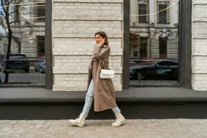 Confident short haired woman wearing  casual   beige color coat, white textured leather shoulder bag, walking in street of European city.  Autumn fashion trends.  Full lenght. photo