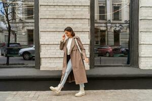 Confident short haired woman wearing  casual   beige color coat, white textured leather shoulder bag, walking in street of European city.  Autumn fashion trends.  Full lenght. photo