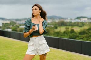 Pretty  brunette woman  drinking coffee outdoor. Elegant accsesorises, leather  bag, stylish crop blouse with puff sleeve. photo