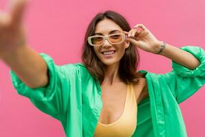 Beautiful graceful woman in  casual outfit posing  on pink background and making self portrait. Wearing  yellow crop top and jeans. Summer trendy look. photo