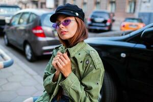 hermosa niña mirando a cámara. cerca arriba retrato de de moda dama en verde chaqueta sentado cerca la carretera exterior. coche estacionamiento antecedentes. foto