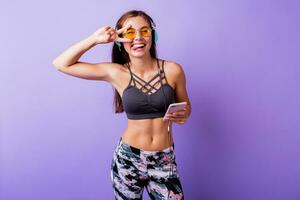 salido deportivo mujer con sincero sonriente posando en elegante activo vestir en estudio. utilizando teléfono inteligente y escuchando música. foto