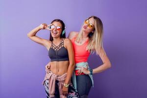 dos bonito muchachas en de moda ropa deportiva posando en púrpura antecedentes en estudio. morena y rubio mujer en pie juntos. aptitud y activo Moda concepto. foto