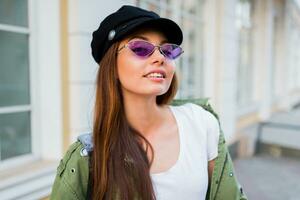 contento morena mujer posando al aire libre . elegante sombrero, Gafas de sol y verde chaqueta. foto