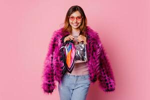 Wonderful girl in fancy pink faux fur short coat holding silver hand bag and looking forward.  Fashion studio shot. Winter season. Shopping concept. photo