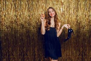 Carefree gorgeous blonde woman  holding glass of  cocktail and shoes in hand. New year party time. Golden sparkly background. photo