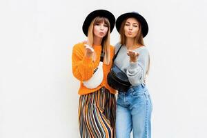 Best friends  embracing , couple of  stylish girls posing on white background. Fall season. Wearing stylish orange knitted sweater, black hat , bum bag. Friends spending great time together. photo