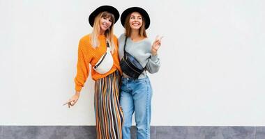 Outdoor lifestyle portrait of couple of young women having fun together.  Windy hairs. Two girls fooling around and dancing on white background. Stylish bum bags, similar black hats. photo