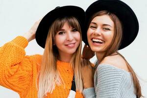 Two carefree girls sending Air kiss to camera , standing over white background. Wearing similar hats and knitted sweaters. photo