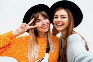 amistad, felicidad y personas concepto. dos sonriente muchachas susurro chisme en blanco antecedentes. naranja suéter, negro similar sombreros. foto