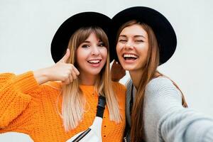 amistad, felicidad y personas concepto. dos sonriente muchachas susurro chisme en blanco antecedentes. naranja suéter, negro similar sombreros. foto