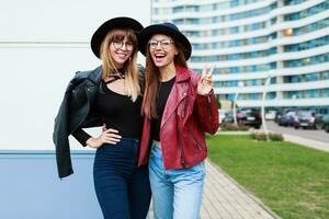 Two carefree smiling women posing  over modern city background. Wearing wool hat , leather jacket and jeans. Friends hugging. Happy emotions . photo