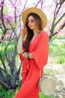 Stylish  bohemian girl posing in coral dress and straw hat near blossom cherry tree in spring park. Holding Bali rattan  straw  bag. photo