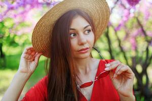 Fresh spring portrait of cute smiling woman in stylish coral dress, in straw hat enjoying sunny day , romantic mood. Flowers background. Candid smile. Weekends  in park. photo