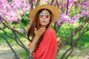 encantador romántico niña enviando aire Beso a cámara en soleado primavera día. rosado floreciente arboles en antecedentes. vistiendo Paja sombrero y coral vestido. foto