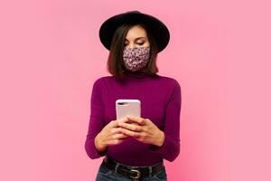 Stylish woman in protective  mask and black hat using mobile phone , standing on pink background. photo