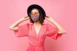 Fashionable woman dressed protective stylish face mask. Wearing black hat and sunglasses. Posing over pink background in studio. photo