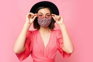 Fashionable woman dressed protective stylish face mask. Wearing black hat and sunglasses. Posing over pink background in studio. photo