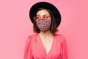 European model dressed protective stylish face mask. Wearing black hat and sunglasses. Posing over pink background in studio. photo