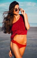 hermosa sexy Delgado mujer en rojo bikini y camiseta con lentes posando en el playa en contra un azul cielo. verano humor. foto