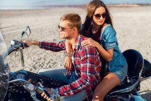 Lifestyle sunny  portrait of young couple riders sitting together on sand beach by motorbike - travel concept. Two people and bike .Fashion woman and man hugging and  smiling. photo