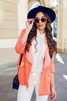 Outdoor fashion portrait of sexy stylish casual woman  in black hat, pink suit ,  white blouse posing on old street background. Spring , fall sunny day. Wavy hairstyle. photo