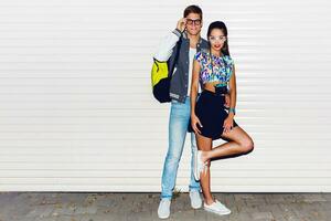 Fashion image of  stylish young  couple in love , tourists , posing near white urban wall. Wearing cool spring clothes, vintage camera,  bright neon back pack , sneakers. photo