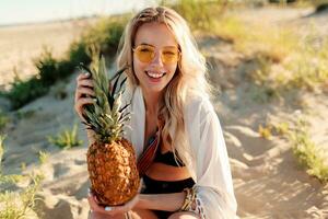 Picture of laughing pretty woman in trendy summer outfit with pineapple relaxing on sunny beach. photo