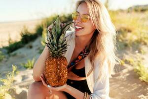 Picture of laughing pretty woman in trendy summer outfit with pineapple relaxing on sunny beach. photo