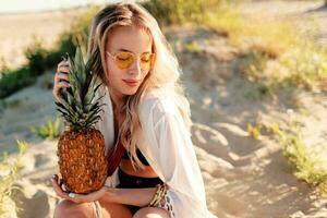 Picture of pretty woman in trendy summer outfit with pineapple relaxing on sunny beach. photo