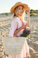 Romantic blonde woman in hat and elegant pink dress on the beach.Holding straw bag and flowers. photo
