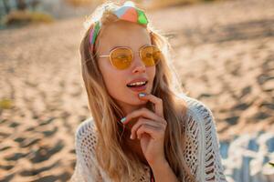 Warm summer portrait of stunning graceful woman enjoying sunset near ocean, sitting on sand. Perfect smile, wearing white boho dress and colorful headband. photo