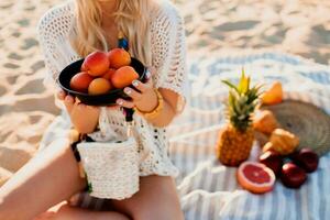 Beautiful blond girl holding plate with peach and relaxing on the evening tropical beach, sunset colors. Sitting on towel. Tropical fruits. Wearing stylish headband and boho dress. Healthy life. photo