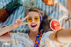 emocionado niña haciendo selfie en playa toalla. boho accesorios. amarillo gafas de sol.arriba vista. foto