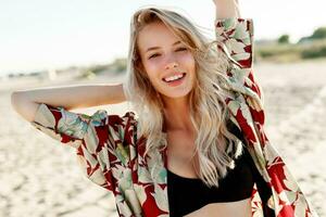 Excited blond woman with perfect smile having fun and looking at camera on sunny white sand beach. photo