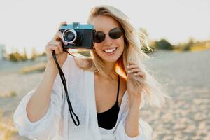 Blonde woman holding retro camera and having fun on warm sunny beach. Sunglasses, white outfit. photo