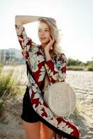 Gorgeous blond woman in trendy beachwear with floral print looking at camera on sunny beach. photo