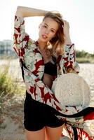Gorgeous blond woman in trendy beachwear with floral print looking at camera on sunny beach. photo
