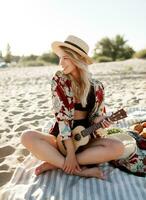 Romantic blonde woman in straw hat sitting on cover on the beach and playing ukulele guitar. Picnic photo