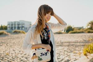 romántico niña con largo pelo caminando en el playa. bohemio estilo, Paja bolso , brillante hacer arriba. foto