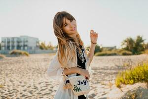 Charming girl with wavy  hairs, dressed in white boho cover up, walking on sunny summer beach. photo