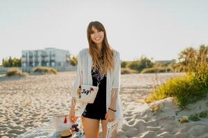 Beautiful woman with wavy hairs, dressed in white boho cover up looks into camera on the beach. photo