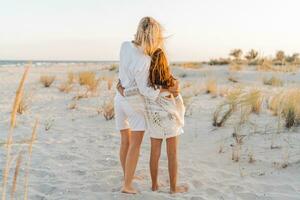 European  mom with cute little daughter posing on sunny beach. Wearing light boho summer clothes. Sunset light. Family vacations concept. photo